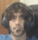 A young man with curly hair wearing a gaming headset makes a concentrated, slightly goofy facial expression.