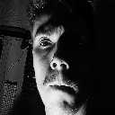 Man with a mustache looking directly at the camera with half of his face obscured by deep shadows.