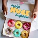 Hands holding an open box of six colorful frosted donuts with text inside the lid.