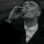 A man in a dark coat smokes a cigarette while looking upward with a serious expression.