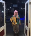 Person with a pink wig and green shirt stands in a doorway holding an acoustic guitar.