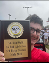 A person wearing glasses and a red shirt holds up a wooden plaque trophy from a 5K race.