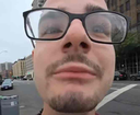 A man with a goatee and glasses stares directly into the camera in an extreme, distorted close-up.
