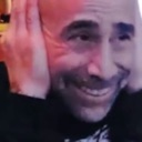 A man with a wide, strained smile presses his hands firmly against his ears.
