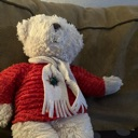 A white teddy bear wearing a red sweater and a white scarf sits on a brown couch.