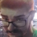 A close-up of a man wearing glasses looking at the camera with a skeptical and judging expression.
