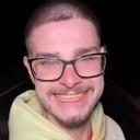A young man with short hair and glasses wearing a yellow hoodie, smiling broadly with a slightly strained expression in a da…