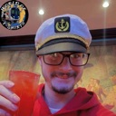 A person wearing a sailor hat and glasses smiles while holding up a glass of red liquid.
