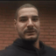 A man with a short beard looking at the camera with a serious and skeptical expression.