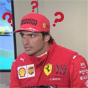 Carlos Sainz wearing a red racing suit and cap with red question marks floating around his head.