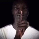Man holding a finger to his lips in a shushing gesture against a dark background.