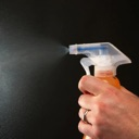 A hand holds a spray bottle and sprays a mist against a dark background.