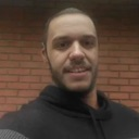 A man with a beard wearing a black hoodie smiles slightly in a casual, grainy selfie.