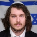 Man with long brown hair and a yarmulke stares directly at the camera with a serious expression in front of an Israeli flag.