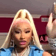 Nicki Minaj with blonde hair and a blue top performing a dismissive hand wave motion.