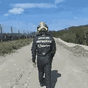 A person wearing a flight suit walks away down a dirt road under a blue sky.