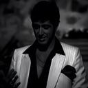 Man in a white suit and dark shirt looking down with a serious expression.