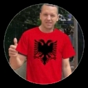 A man wearing a red shirt with a black double-headed eagle emblem gives a thumbs-up gesture.