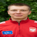 A man with short hair wearing a red athletic jacket with a white eagle emblem on his chest.