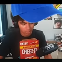 A person wearing a blue cap and headphones holds a red box of crackers against their chest.