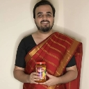 A man with a beard wearing a red shawl over a black shirt holds up a jar of food with a happy expression.