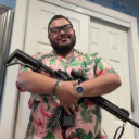 A man wearing a pink floral shirt smiles while holding a rifle across his chest.