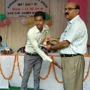 A man in a light shirt hands a trophy to a younger man during an indoor event.