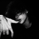 A person with dark hair and glasses partially obscuring their face, posing with a hand near their mouth in a high-contrast b…