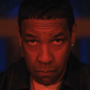 Denzel Washington stares directly forward with a serious expression under intense red lighting.