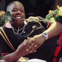 A man reclining while holding a red rose in his teeth with a confident smile.