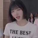 A young woman with short hair wearing a white shirt makes a finger heart gesture toward the camera.