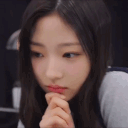 A young woman with dark hair rests her chin on her hand while looking down with a serious, pensive expression.