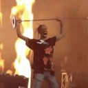 Travis Scott screaming while holding a microphone stand above his head in front of flames.