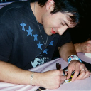 A young man with dark hair wearing a star-patterned shirt focuses intently while signing an autograph.