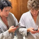 Two men wearing gray bathrobes looking down at their smartphones with serious expressions.
