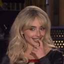 Sabrina Carpenter smiling coyly with her finger touching her lower lip.