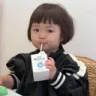 A young child with a bob haircut looking directly at the camera while sipping from a small milk carton with a straw.
