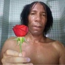 A man holding a single red rose toward the camera with a serious expression.