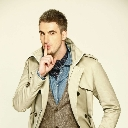 Man in a trench coat holding a finger to his lips in a shushing gesture.