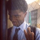 A young man with a smirk peers through a wire-reinforced window with his hand pressed against the glass.