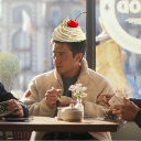 Simu Liu sits at a table with a cupcake balanced on top of his head.