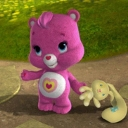A pink Care Bear standing in a grassy area holding a small yellow toy.