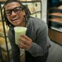 A man wearing glasses smiles broadly while holding up a green drink in a plastic cup.