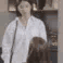 A woman in a white shirt stands in a kitchen with a blank, unimpressed expression on her face.