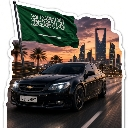 A black car driving on a road with the Saudi Arabian flag waving above and the Kingdom Centre skyscraper in the background.