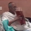 A man sits comfortably while holding and sipping from an extremely large, tall glass filled with a dark beverage.