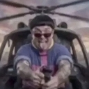 A person wearing a beanie and sunglasses sticks their tongue out while piloting a helicopter.