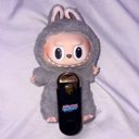 A gray plush creature with a wide, toothy grin peeking out from behind a black electronic device.