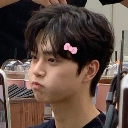 A young man with dark hair and a small pink bow clip in his hair making a pouting face with puffed cheeks.
