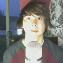 Young man with messy hair looking directly at the camera with a blank expression while positioned behind a microphone.